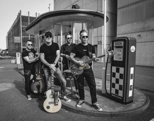 Frankfort Special Band posiert mit Instrumenten neben einer alten Benzinpumpe in schwarz-weiß.