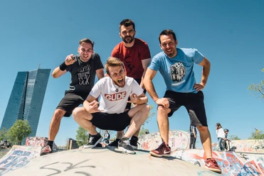 Alex im Westerland Vier Männer posieren fröhlich in einem Skatepark bei Sonnenschein, mit modernen Gebäuden im Hintergrund.