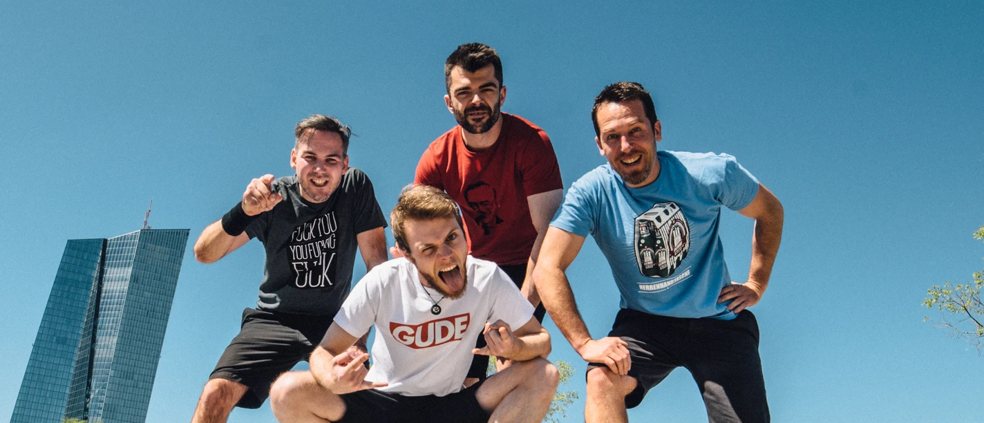 Alex im Westerland Vier Männer posieren fröhlich in einem Skatepark bei Sonnenschein, mit modernen Gebäuden im Hintergrund.