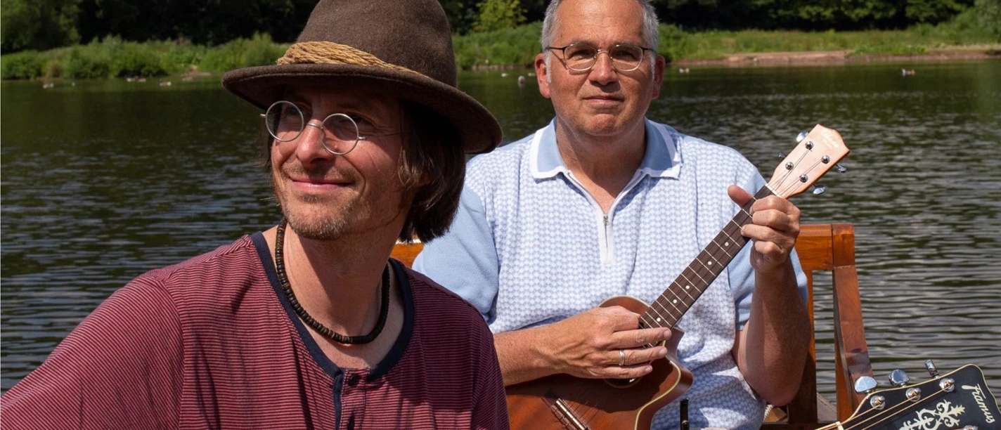 KULT-Net Bühne - DER RAMSCHLADEN Zwei Musiker von Der Ramschladen spielen Gitarre am See.