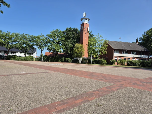 Gepflasterter Parkplatz am Marktplatz mit Bäumen, Wohnhäusern und hohem Backsteinturm.