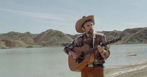 Shota Adamashvili mit Cowboyhut und Gitarre vor See und kargen Hügeln