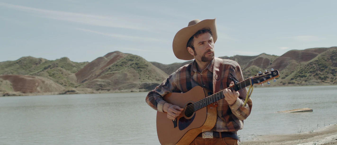 Shota Adamashvili mit Cowboyhut und Gitarre vor See und kargen Hügeln