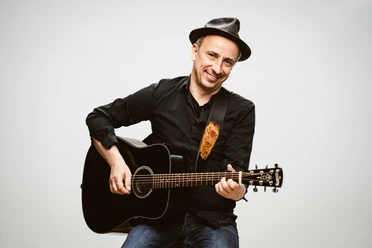 Stefan Knittler lächelt mit schwarzer Gitarre und Hut vor hellem Hintergrund