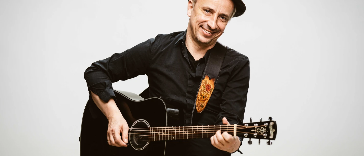 Stefan Knittler lächelt mit schwarzer Gitarre und Hut vor hellem Hintergrund