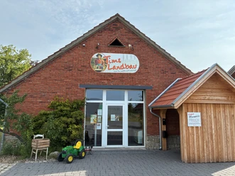 Rotes Ziegelsteinhaus mit großer Eingangtür. Darüber ein Werbeschild mit der Aufschrift: Tims Landbau. Im Vordergrund ist ein kleiner Spielzeugtrekker zu sehen.