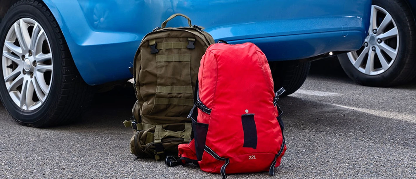 Two backpacks are resting on the ground next to a blue car in a parking lot during daytime. Travel concept