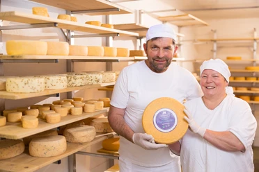 Heike und Friedrich Dörmann Halten ein Laib Käse, im Hintergrund Regale mit zahlreichen großen und kleinen Käselaiben