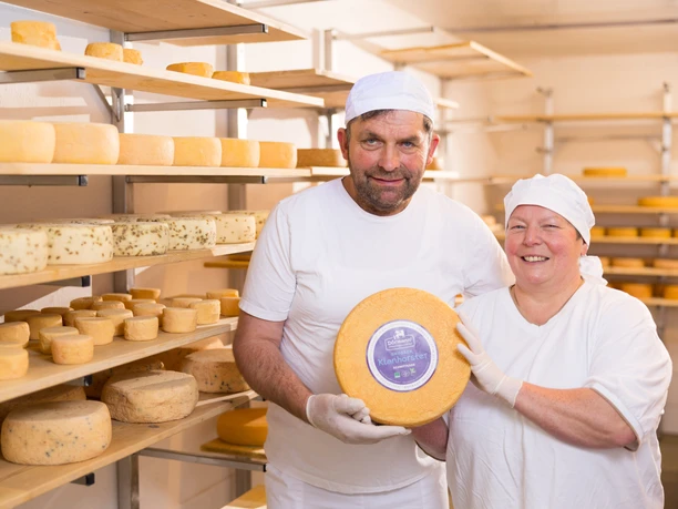 Heike und Friedrich Dörmann Halten ein Laib Käse, im Hintergrund Regale mit zahlreichen großen und kleinen Käselaiben