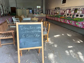 Innenraum einer Deele. Im Hintergrund ist ein Verkaufstresen zu sehen, rechts eine lange Reihe von Kisten mit Gemüse, im Vordergrund eine Tafel mit der Aufschrift Tomaten zum selber pflücken für 6 Euro das Kilo, dahinter Holzbänke und Tische.