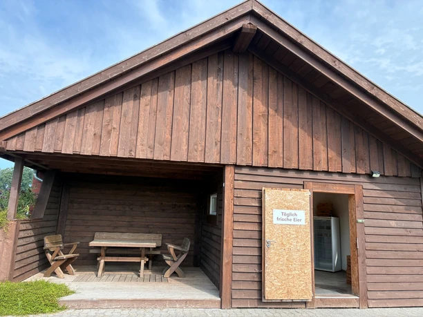 Braune Holzhütte mit Spitzdach. Links ein überdachter Sitzplatz, rechts die offene Eingangstür.