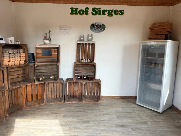 Raum mit Holzkisten stehen mit Eiern, Honig und Marmeldaden. An der weißen Wand steht in grüner Schrift Hof Sirges.