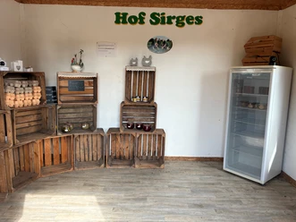 Raum mit Holzkisten stehen mit Eiern, Honig und Marmeldaden. An der weißen Wand steht in grüner Schrift Hof Sirges.