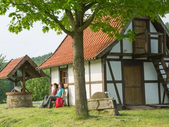 Fachwerkhaus mit Brunnen, im Grünen gelegen; zwei Personen sitzen auf einer Bank vor dem Haus.Half-timbered house with a fountain, surrounded by greenery; two people sitting on a bench in front of the house.Bindingsværkshus med springvand, omgivet af grønne områder; to personer sidder på en bænk foran huset.Vakwerkhuis met fontein, omgeven door groen; twee mensen zitten op een bankje voor het huis.