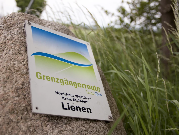Radeln auf der Grenzgängerroute Schild der Grenzgängerroute Teuto-Ems in Lienen, umgeben von hohen Gräsern.Sign for the Teuto-Ems cross-border route in Lienen, surrounded by tall grasses.Skilt til den grænseoverskridende rute Teuto-Ems i Lienen, omgivet af højt græs.Bord voor de grensoverschrijdende route Teuto-Ems in Lienen, omgeven door hoog gras.
