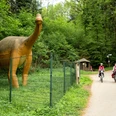 Dinosaurier bei den Dinosaurierspuren bei Barkhausen Zwei Radfahrer auf einem Waldweg, neben einer großen Dinosaurier-Statue im Grünen.