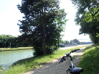 Radeln auf der OC2 - Rund um Schwagstorf Fahrradweg am Ufer eines Kanals mit Brücke im Hintergrund, umgeben von Bäumen.