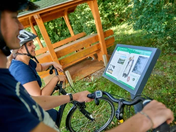 Hörstation auf der Ackerschnacker Tour Zwei Radfahrer betrachten eine Infotafel neben einem überdachten Holzunterstand im Grünen.Two cyclists look at an information board next to a covered wooden shelter in the countryside.To cyklister kigger på en informationstavle ved siden af et overdækket træskur på landet.Twee fietsers kijken naar een informatiebord naast een overdekt houten afdak op het platteland.