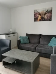 Neanderglück am Kaldenberg Modern eingerichteter Wohnraum mit graufarbigem Sofa, zwei hellblauen Sofakissen, einem Tisch und zwei Sesseln. Ein schönes Foto von einem Hochlandrind mit Hörnern und langem Fell hängt über dem Sofa. Eine hüfthohes weißes Schränkchen mit zwei Türen steht in Blickrichtung links neben dem Sofa.