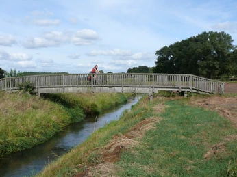 Radeln auf dem Brückenradweg Holzbrücke über einen kleinen Fluss, flankiert von grünen Wiesen unter einem wolkigen Himmel.Wooden bridge over a small river, flanked by green meadows under a cloudy sky.Træbro over en lille flod, flankeret af grønne enge under en overskyet himmel.Houten brug over een kleine rivier, geflankeerd door groene weiden onder een bewolkte hemel.