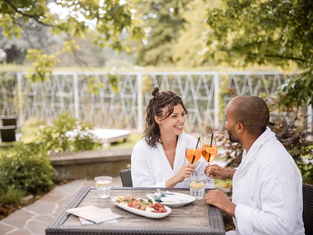 Zwei Gäste im Bademantel stoßen auf der Gartenterrasse der Claudius Therme an