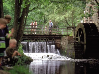 Radeln auf der Niedersäschischen Mühlentour Kinder spielen am Bach, während Erwachsene auf einer Brücke neben einem alten Wasserrad stehen.