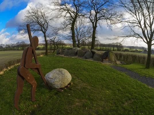 Radeln auf der Radroute der Megalithkultur Metallskulptur eines Menschen, der einen großen Stein inmitten einer grünen Wiese rollt.Metal sculpture of a man rolling a large stone in the middle of a green meadow.Metalskulptur af en mand, der ruller en stor sten midt på en grøn eng.Metalen beeld van een man die een grote steen rolt in het midden van een groene weide.