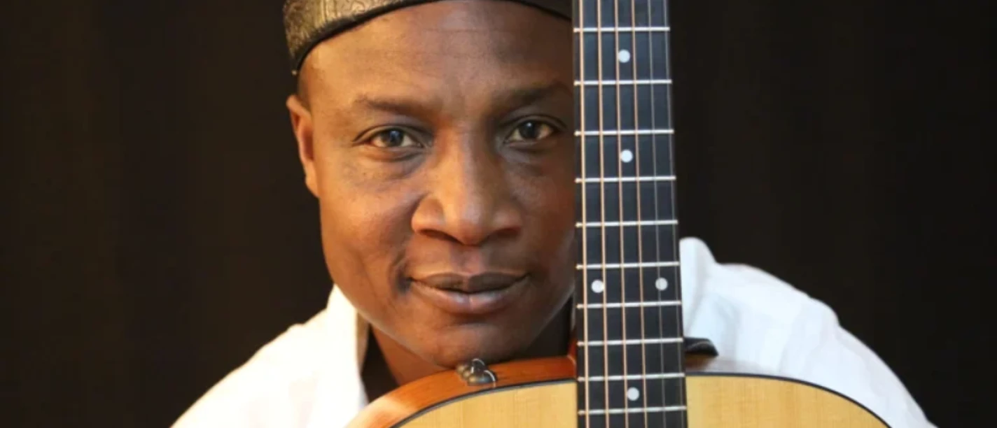 Musiker Adjiri Odametey mit Lederkappe und weißem Hemd hinter einer Akustikgitarre.