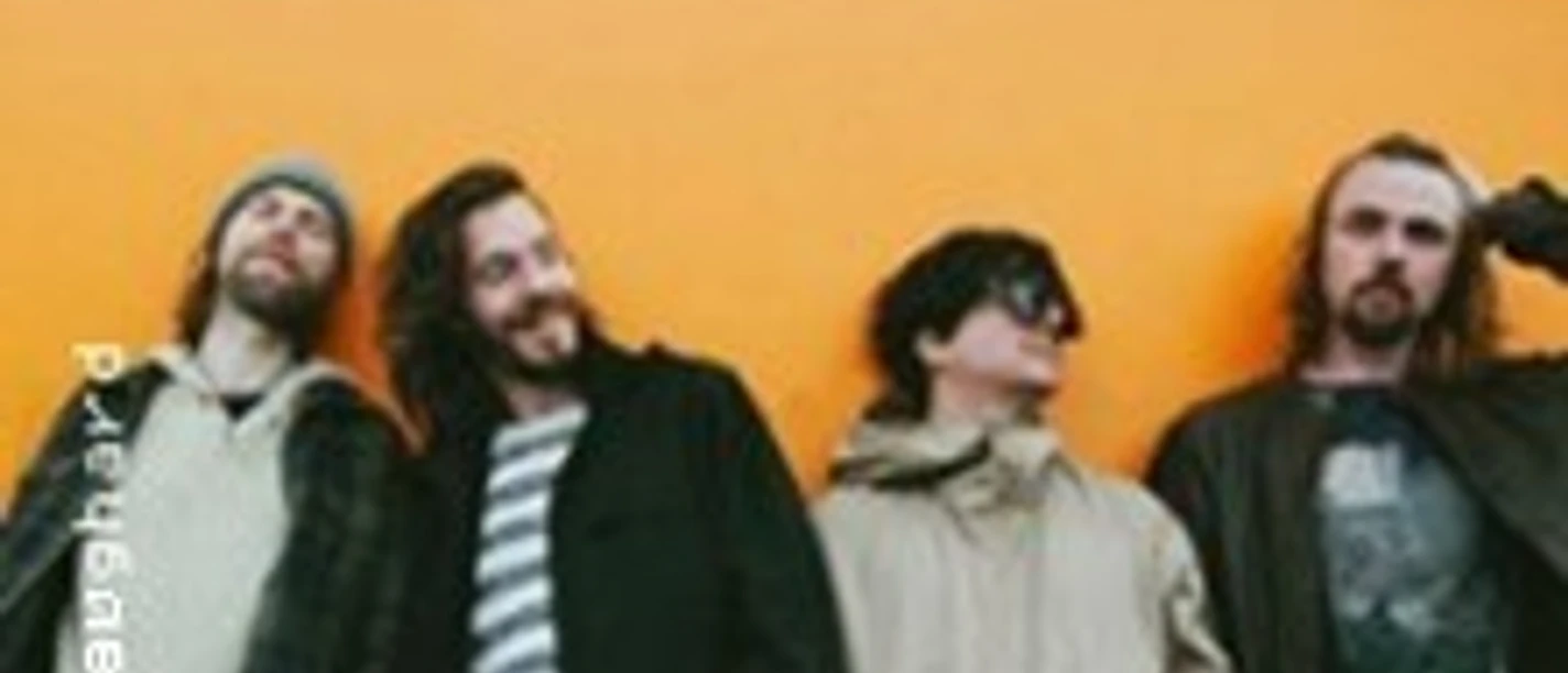 Vier DenManTau-Musiker vor orangefarbener Wand in entspannter Bandpose