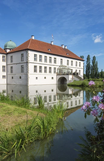 Radeln auf der NST -Tour Weißes Wasserschloss mit roten Dächern, umgeben von einem Wassergraben unter blauem Himmel.White moated castle with red roofs, surrounded by a moat under a blue sky.Hvid vandborg med røde tage, omgivet af en voldgrav under en blå himmel.Witte waterburcht met rode daken, omgeven door een slotgracht onder een blauwe hemel.