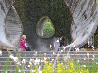 Radeln auf der NST -Tour Zwei Personen sitzen in einer modernen, offenen Holzkonstruktion, umgeben von Wildblumen.Two people sit in a modern, open wooden structure surrounded by wild flowers.To personer sidder i en moderne, åben trækonstruktion omgivet af vilde blomster.Twee mensen zitten in een moderne, open houten constructie omringd door wilde bloemen.