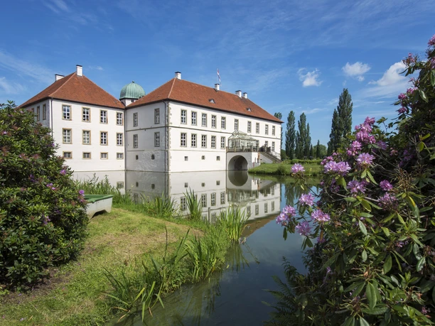 Radeln auf der BE 3 - Rund um Bad Essen Barockschloss mit rotem Dach spiegelt sich in stiller Wassergrabenlandschaft bei blauem Himmel.