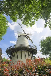 Unterwegs auf derOsning-Route Weißer Mühlenturm umgeben von blühenden Pflanzen und grünem Laub unter blauem Himmel.