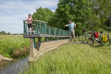 An der Bifurkation Twee fietsers op een brug in een groen landschap met een rivier en bomen op de achtergrond.