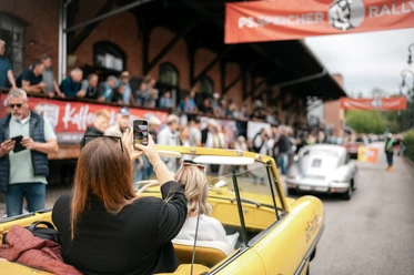 PS.SPEICHER Rallye 2026 Frau fotografiert aus gelbem Oldtimer-Cabriolet die PS.SPEICHER Rallye mit Zuschauern.