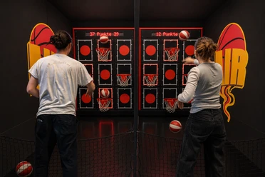 Two people are throwing basketballs at illuminated baskets at a game show station