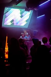 DJs vor Publikum im Club Z, umgeben von violettem Licht und grafischen Projektionen