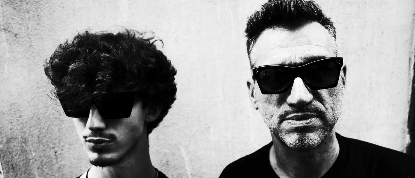Two men wearing sunglasses in front of a wall; black-and-white photo.