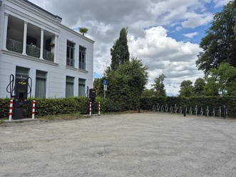 Parkplatz mit EWE-Go-Ladesäulen, Fahrradständern und weißem Gebäude unter Wolkenhimmel.