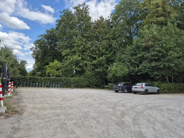Waldparkplatz mit E-Ladesäule, Fahrradständern und geparkten Autos vor dichtem Baumbestand.