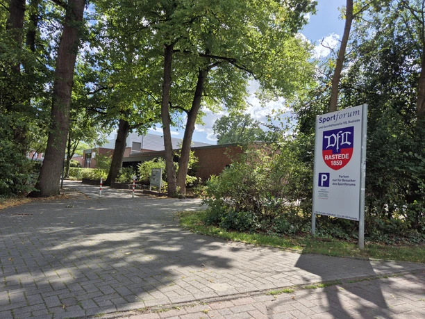 Eingang zum Sportforum des VfL Rastede mit Hinweisschild unter hohen Bäumen.