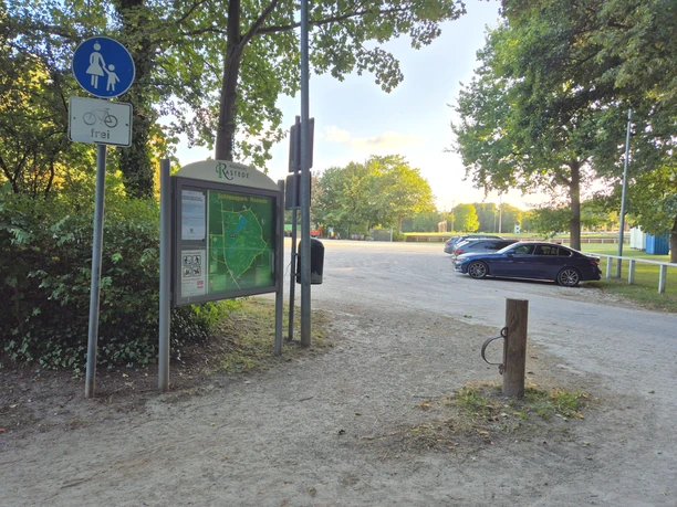 Parkplatz am Ellernteich mit Infotafel, Fußwegschild und parkenden Autos unter Bäumen.