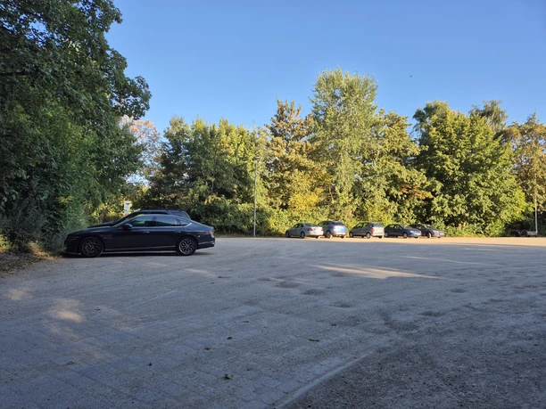 Schotterparkplatz am Ellernteich mit parkenden Autos vor dichtem Baumbestand.