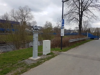 Parkplatz mit Beschilderung und Stromsäule am Flussufer nahe einer blauen Brücke