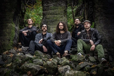 Five musicians seated among stone walls in greenery
