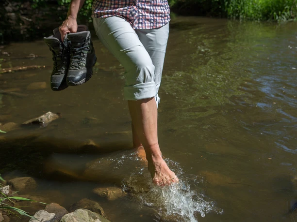 Wassertreten in der Nette Barefoot person wading through a shallow stream and holding hiking boots in one hand.