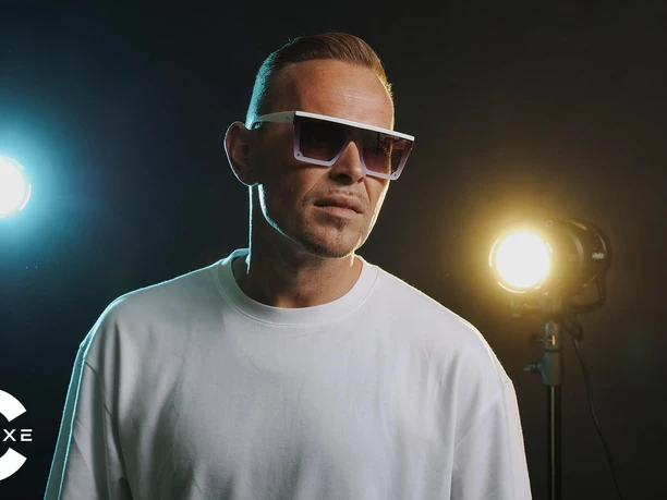 Imagefoto: C-Deluxe trägt Sonnenbrille und weißes Shirt und steht mit lässiger Körperhaltung vor einem dunklen Hintergrund mit einem blauen und gelben Scheinwerfer.