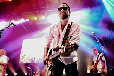 Mann mit Sonnenbrille spielt E-Gitarre auf einer beleuchteten Bühne.