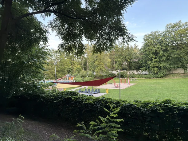 A sunbathing area at the Wiehe outdoor pool with a shade sail, trees, and a play area.