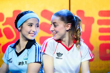 Two female soccer players in jerseys smile at each other against a yellow-and-red backdrop
